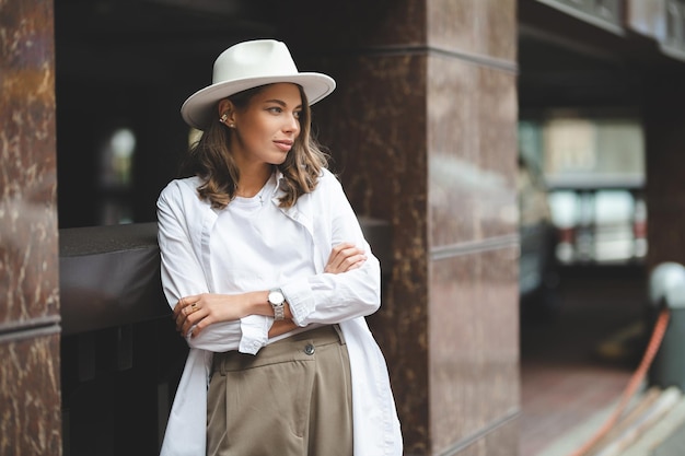 Chica elegante con pantalones blancos y blusa blanca de algodón posando contra la pared