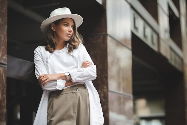 Chica elegante con pantalones blancos y blusa blanca de algodón posando contra la pared