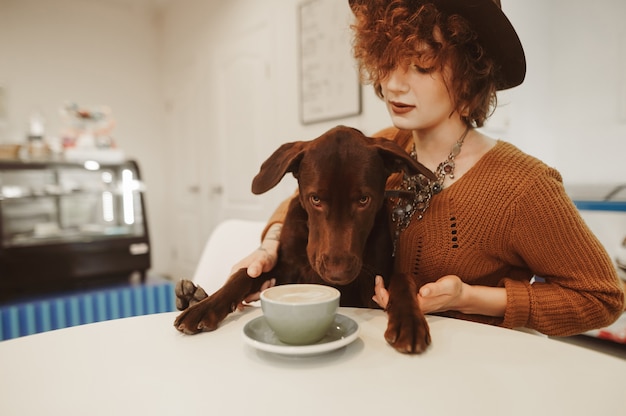 Chica elegante con pelo rizado y sombrero se sienta en la mesa junto a la taza de café con cachorro en las manos