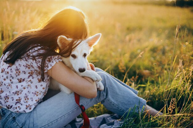 Foto chica elegante relajándose con su adorable cachorro en un picnic amigo leal mujer feliz abrazando a un lindo cachorro blanco en el prado de verano a la luz del atardecer auténtico momento hermoso