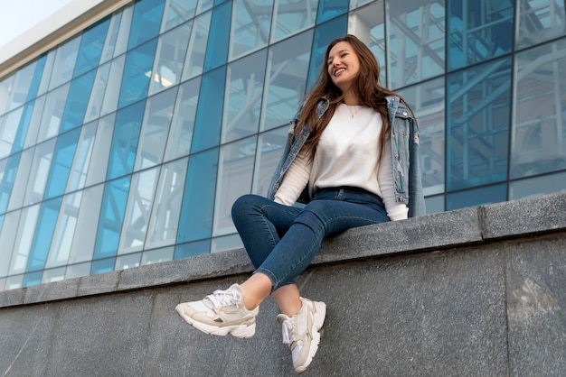 Chica elegante riendo descansando en la calle de la ciudad de verano Hermosa chica estudiante sentada cerca de un edificio moderno