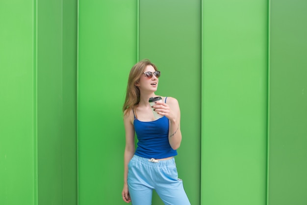 Chica elegante en ropa de calle se encuentra en el fondo de una pared verde con un vaso de café en sus manos. Calle retrato de una mujer posando en el contexto de una pared verde