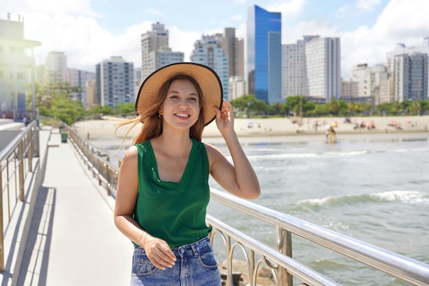 Chica elegante con sombrero caminando por el paseo marítimo en un día ventoso con rascacielos en el fondo Santos Brasil