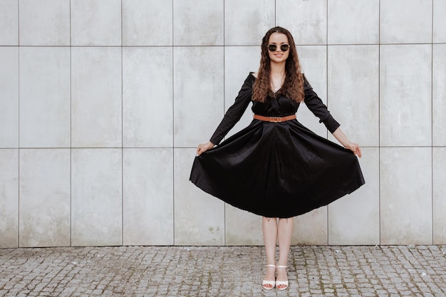 Chica elegante con un vestido negro y gafas de sol cerca de la pared gris. Foto de mujer con gafas. Mujer joven, peinado rizado, sonriendo cerca de una pared en la calle. Chica con ropa de vestir negra.