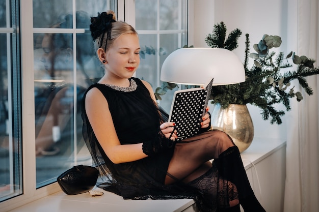 Una chica elegante con un vestido negro se sienta en el alféizar de la ventana con un libro en las manos.