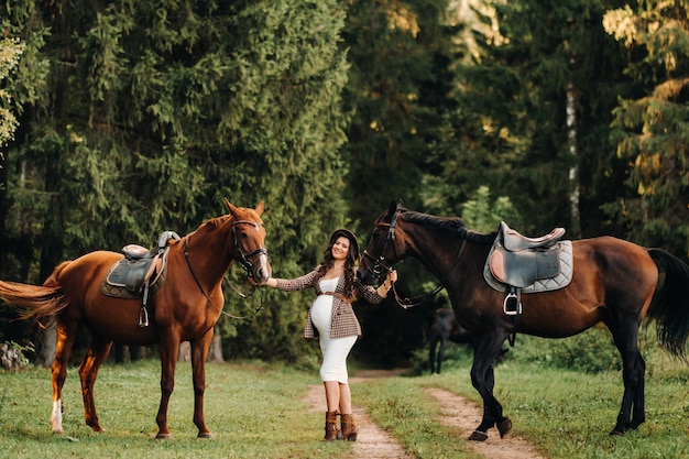 Chica embarazada con una gran barriga en un sombrero junto a caballos en el bosque en la naturaleza. Chica elegante con ropa blanca y una chaqueta marrón.