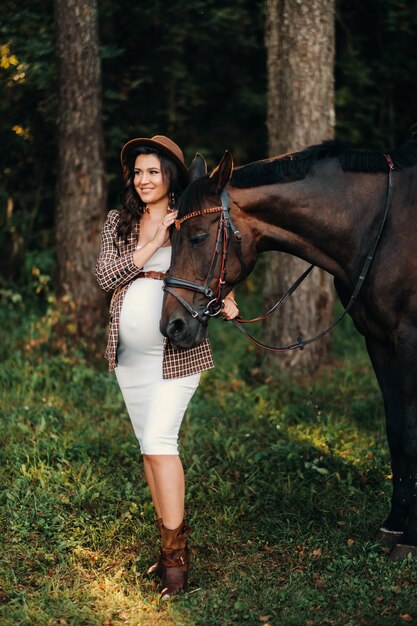 Chica embarazada con una gran barriga en un sombrero junto a caballos en el bosque en la naturaleza. Chica elegante con ropa blanca y una chaqueta marrón.