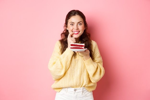 chica emocionada pensando en comer un delicioso pastel, mordiendo el dedo por la tentación y mirando pensativo a la cámara, piensa en las calorías del postre, pared rosa.