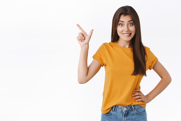 Foto chica emocionada quiere probar algo tentador apuntando a la esquina superior izquierda con aspiración y emoción mordiéndose el labio ansiosa por probar un nuevo producto mirando la cámara de pie fondo blanco quiere espacio de copia fresco