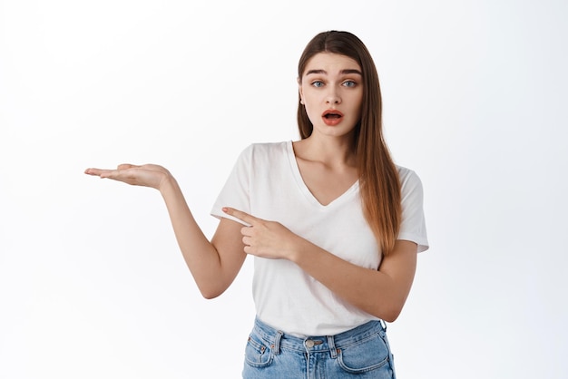 La chica emocionada señala su mano abierta con un espacio vacío para la colocación del producto que muestra el artículo para comprar con la cara intrigada haciendo preguntas sobre la promoción en el fondo blanco de la pantalla