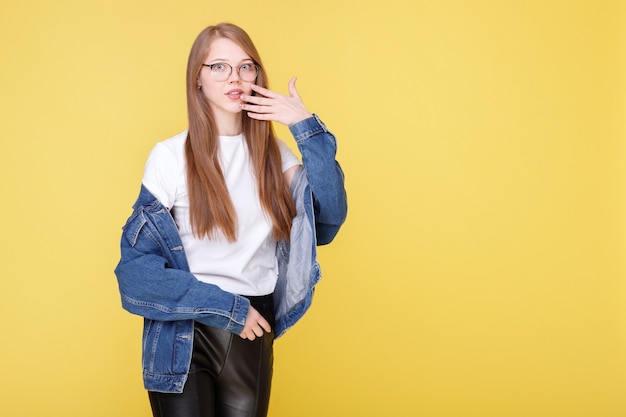 Chica emocional con gafas sorprendida y se cubre la boca con la mano sobre una pared amarilla