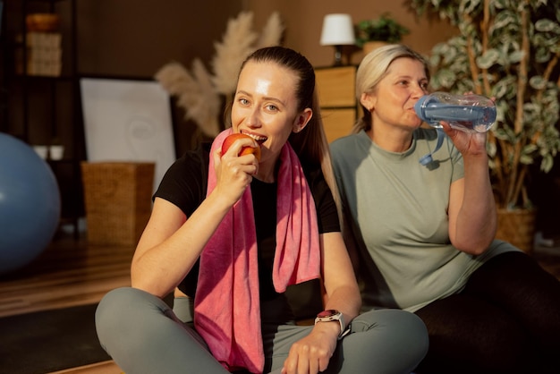 Chica encantada mordiendo manzana madura después de hacer ejercicio con madre anciana Mujer mediana bebe agua