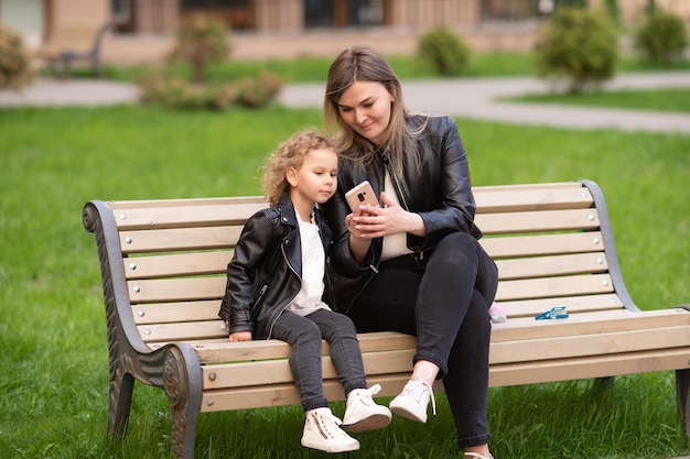 Una chica encantadora con una chaqueta negra está sentada en un banco con su madre y mirando el teléfono.