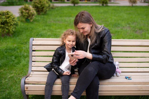 Una chica encantadora con una chaqueta negra está sentada en un banco con su madre y mirando el teléfono.