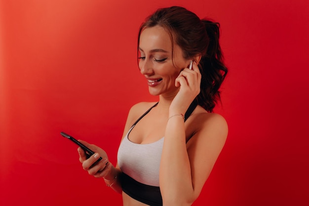 Una chica encantadora y emocionada con cabello oscuro ondulado lleva una camiseta deportiva, escucha música con auriculares y sostiene un teléfono inteligente antes de entrenar