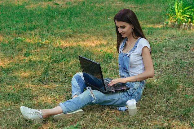 Una chica encantadora se sienta en el parque sobre un césped y trabaja con una laptop