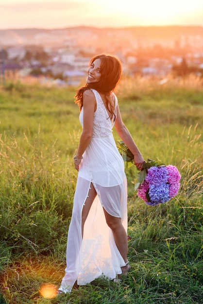 Una chica encantadora con un vestido blanco con un ramo de flores de colores tiernos camina en el césped al atardecer.