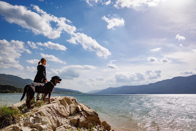 Chica se encuentra cerca del perro de raza Rottweiler en la orilla cerca del lago contra el telón de fondo de la cordillera cubierta de bosque