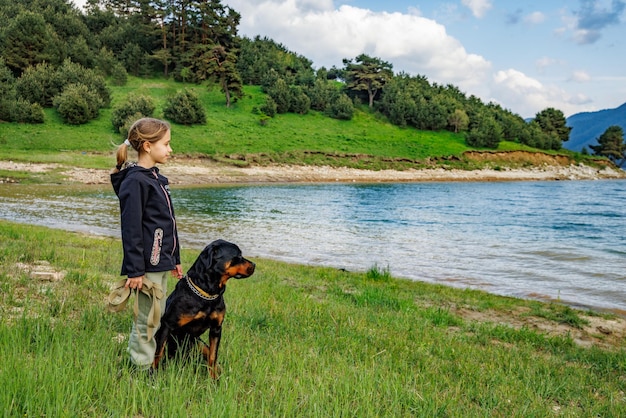 Chica se encuentra cerca del perro de raza Rottweiler en el prado junto al lago contra el valle montañoso con bosques de abetos