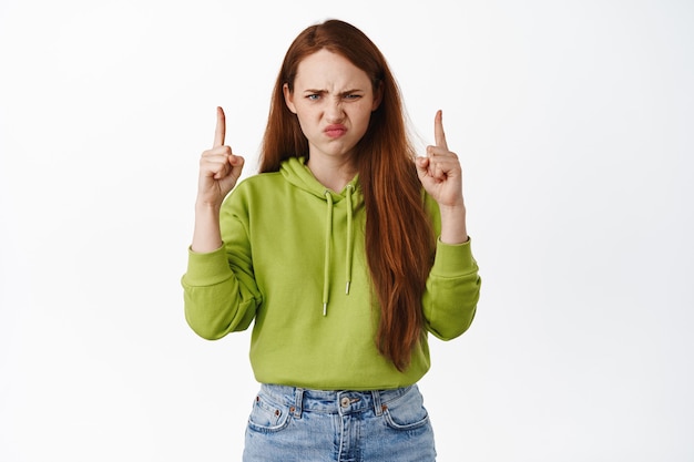 chica enfurruñada con el pelo rojo, haciendo muecas y frunciendo el ceño enojada, molesta por algo molesto en blanco.