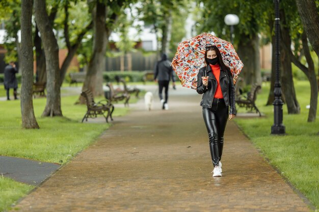 Chica enmascarada está caminando por la calle. Chica con una máscara protectora camina en el parque con un paraguas bajo la lluvia. Infección por coronavirus COVID-19