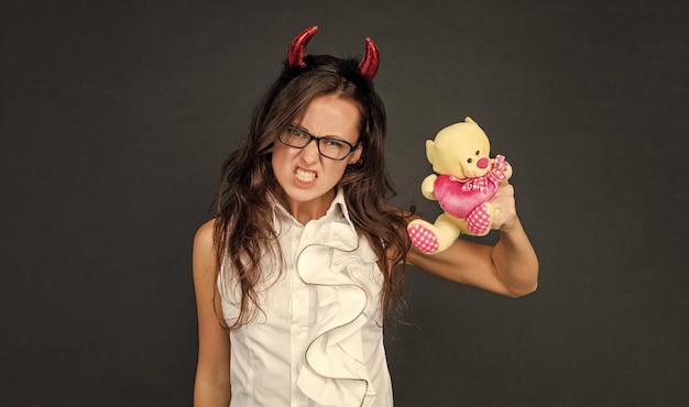 Chica enojada con cuernos de diablo rojo hace cara de miedo sosteniendo el oso de San Valentín fondo oscuro Halloween