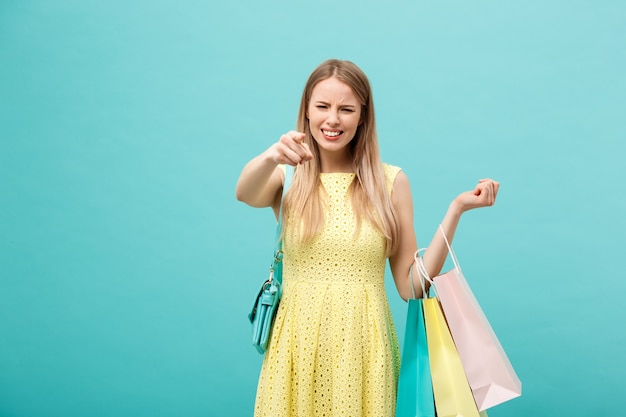 Chica enojada en vestido aislado sobre fondo azul. Sosteniendo la bolsa de papel de la compra para llevar y señalar con el dedo.