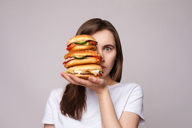 Chica con una enorme hamburguesa en la mano. Retrato de estudio de una joven mujer morena con camiseta blanca con enormes hamburguesas en su mano mirando sorprendido o sorprendido