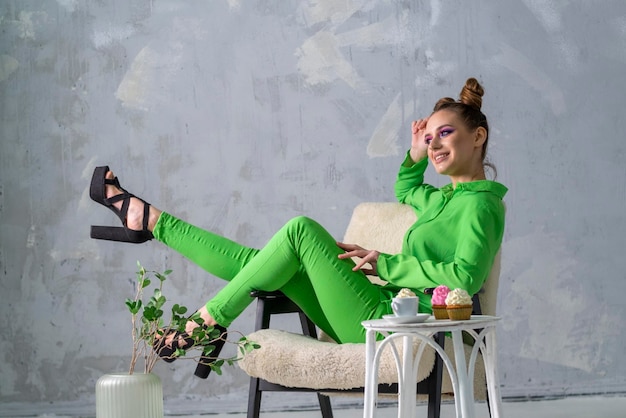 Chica de ensueño posa en silla sobre fondo de pared gris Mujer joven en traje verde sabe cupcakes con crema y café