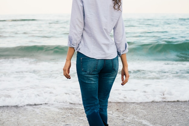 Chica esbelta en camisa y jeans caminando por la playa. vista desde la parte posterior