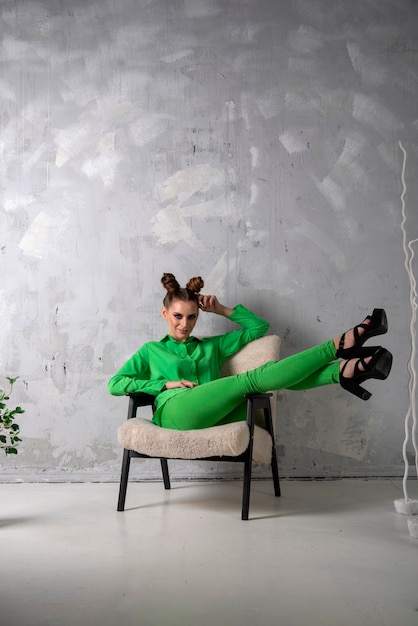 Chica esbelta sexy con elegante traje verde y tacones se sienta en una silla sobre fondo de pared gris en el estudio Marco vertical