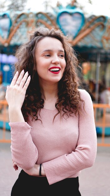 Chica escalofriante en el parque de atracciones en la mañana del fin de semana. Modelo femenino de buen humor de risa con pie de pelo rizado cerca del carrusel.