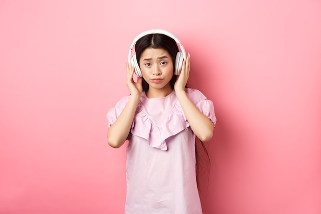 Chica escéptica que no se divierte con la canción, escucha música en auriculares y frunce el ceño disgustada, de pie contra el fondo rosa.