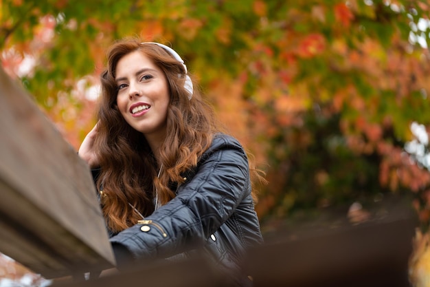 Chica escuchando música con auriculares blancos en un banco del parque en otoño con árboles coloridos en el fondo