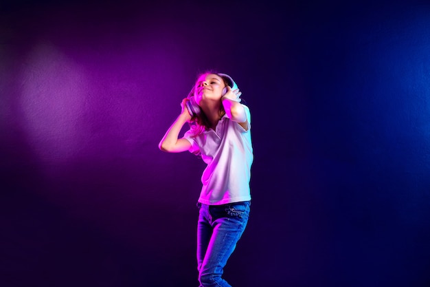 Chica escuchando música en auriculares en colores oscuros. Niño lindo que disfruta de música de baile feliz, ojos cerrados y sonrisa posando
