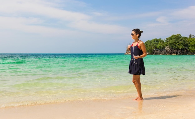 Chica escuchando música en la playa frente al mar mientras sostiene un móvil conectado a los auriculares