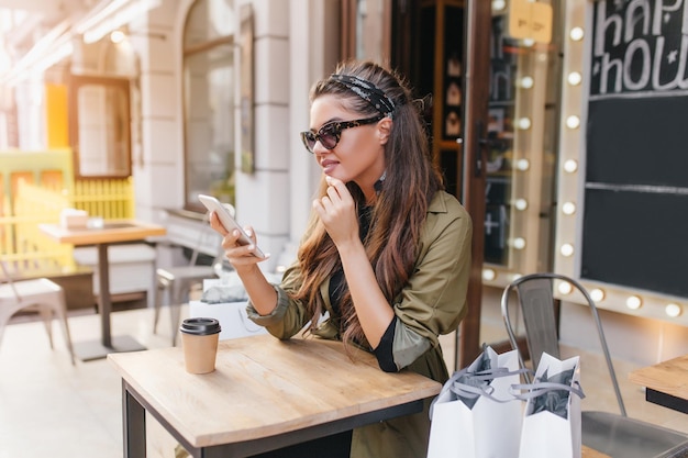 Chica espectacular con mensaje de texto de piel bronceada en el teléfono inteligente mientras se relaja después de ir de compras. Foto exterior de dama bronceada fashionista sentada en la cafetería con compras.