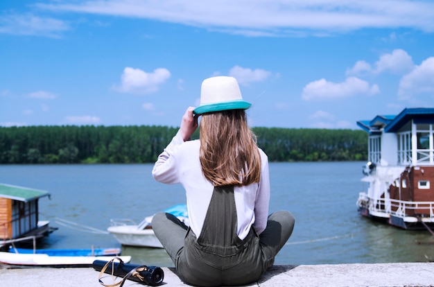 Chica esperando el verano junto al río. Chica de vuelta sentada en la orilla del río y mirando en la isla del bosque en el agua azul.