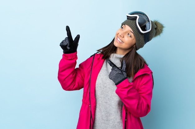 Chica de esquiador de raza mixta con gafas de snowboard en azul aislado señalando con el dedo índice una gran idea