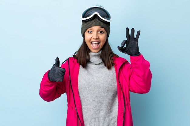 Chica de esquiador de raza mixta con gafas de snowboard sobre azul aislado mostrando signo ok y pulgar arriba gesto