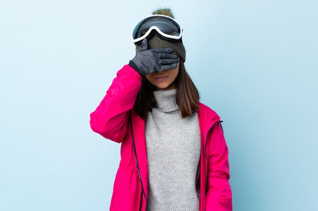 Chica esquiador de raza mixta con gafas de snowboard sobre azul aislado que cubre los ojos con las manos. No quiero ver algo.