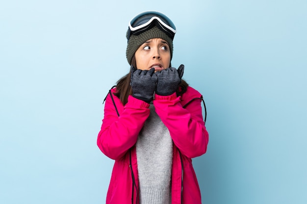 Chica esquiador de raza mixta con gafas de snowboard sobre pared azul aislada nerviosa y asustada poniendo las manos en la boca.