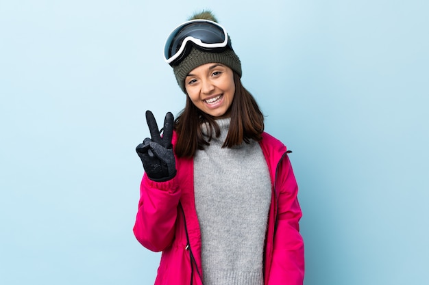 Chica de esquiador de raza mixta con gafas de snowboard sobre pared azul aislado sonriendo y mostrando el signo de la victoria