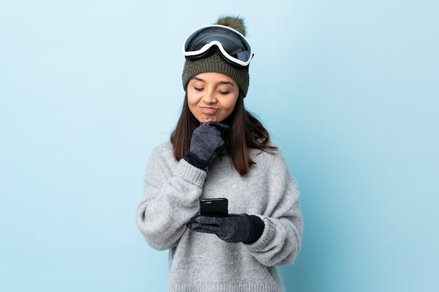 Chica de esquiador de raza mixta con gafas de snowboard sobre pared azul pensando y enviando un mensaje.