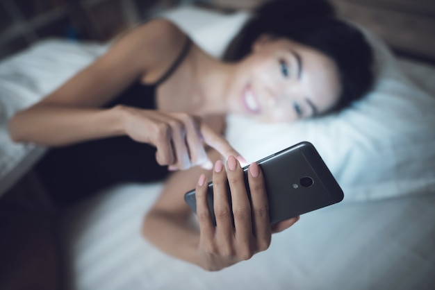 Chica está en un cuarto oscuro en la cama. Ella mira el teléfono inteligente.