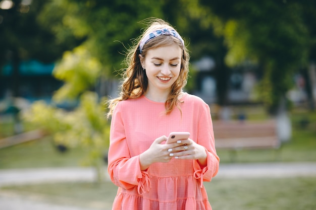 Chica se está riendo y leyendo algo en su teléfono inteligente. Alegre joven comunicarse en teléfono inteligente mientras camina en el parque