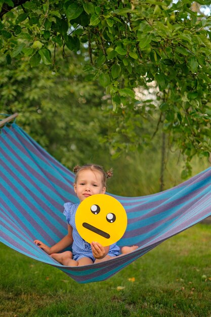 La chica está sentada en una hamaca en el jardín sosteniendo la cara de un emoticon triste y lloroso