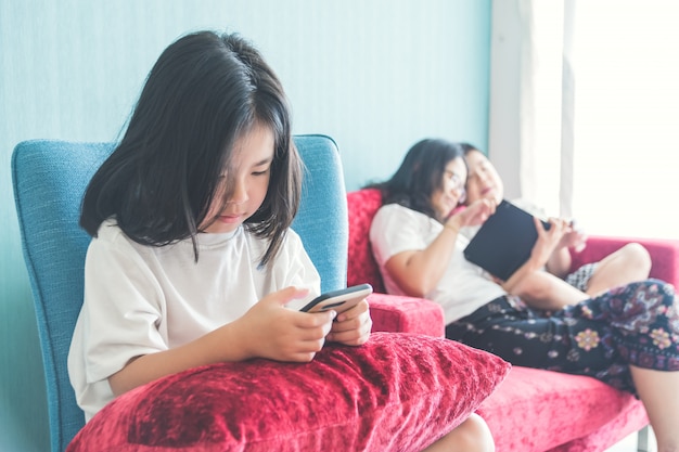 Chica está usando un teléfono inteligente en la silla Madre disfrutando con su hermano en el sofá en casa