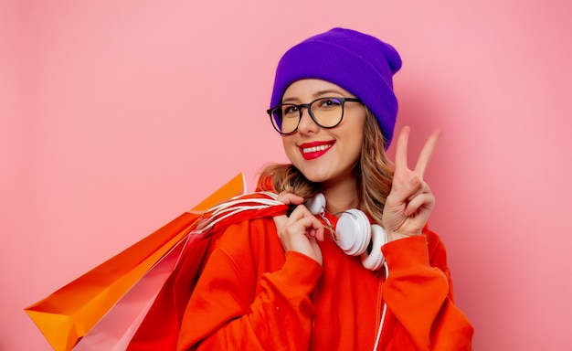 Chica de estilo con capucha naranja y sombrero morado con bolsas de compras en la pared rosa