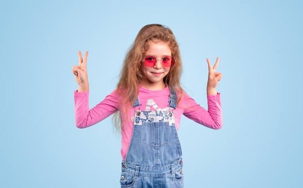 Chica con estilo en denim general y gafas de sol de color rosa de moda que muestran el gesto de dos dedos y mirando a la cámara contra el fondo azul
